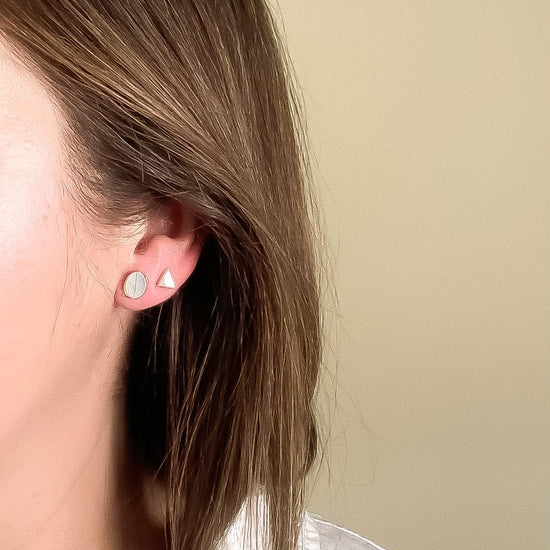 Close-up of a person wearing mother of pearl oval and triangle stud earrings with a beige background