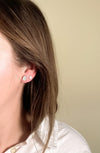 Close-up of a person wearing mother of pearl oval and triangle stud earrings with a beige background