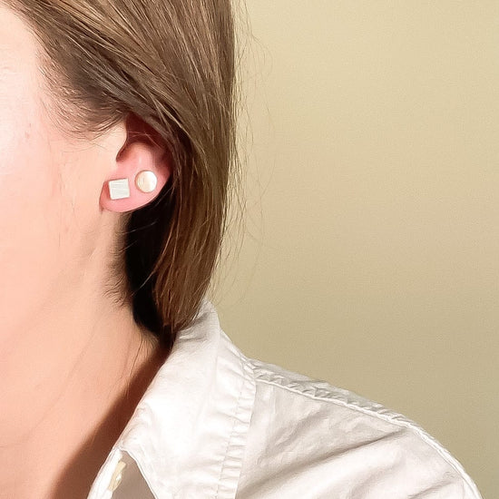 Close-up of a person wearing Mother of Pearl circle and square stud earrings with a plain background