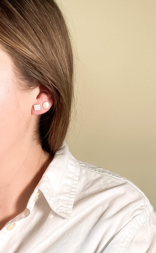 Close-up of a person wearing Mother of Pearl circle and square stud earrings with a plain background